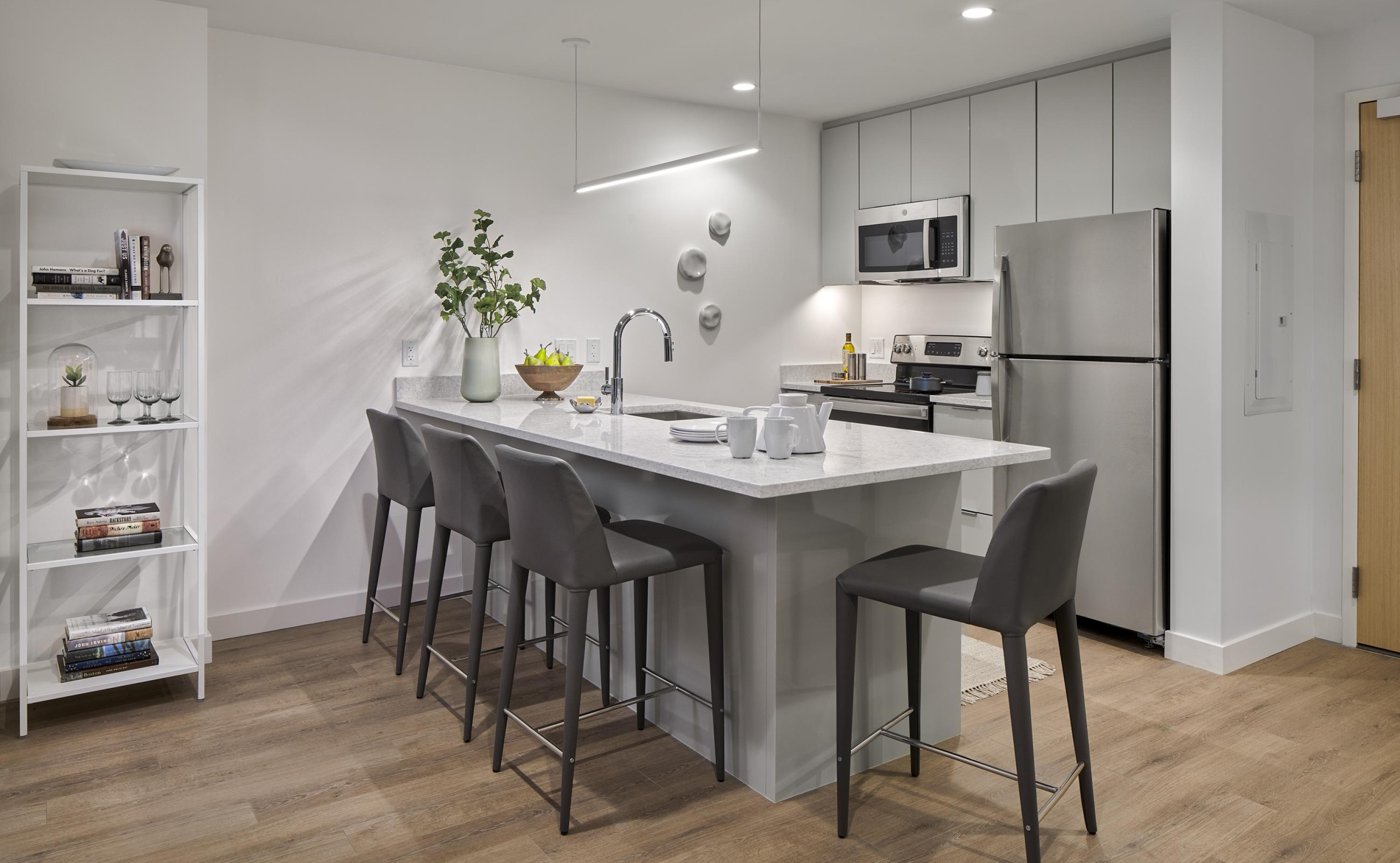 the kitchen with pendant lighting overhead, kitchen island with sink, and stainless steel appliances including fridge, stove, and microwave with grey cabinetry and hard flooring