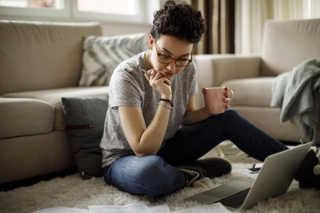 Young woman in glasses holding a pen in one hand and a mug in her other hand, is sitting on the ground doing work.  