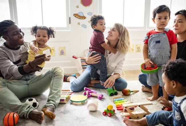 Three parents sitting by four young toddlers on the ground surrounded by toys.