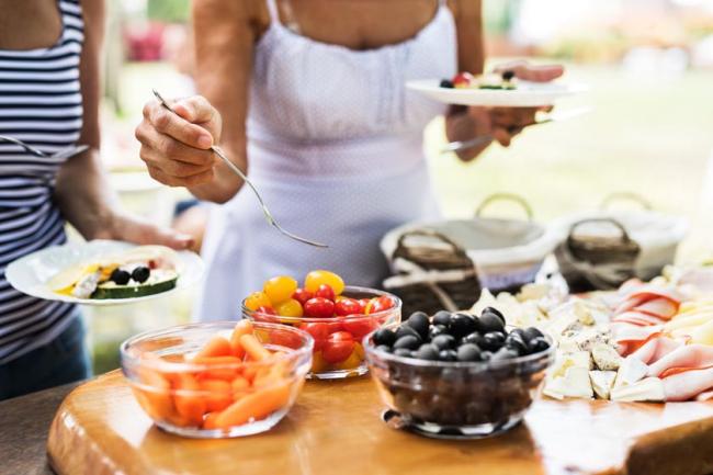 A woman holding a fork above three glass bowls filled with carrots, small tomatoes, and blueberries at an outdoor gathering.