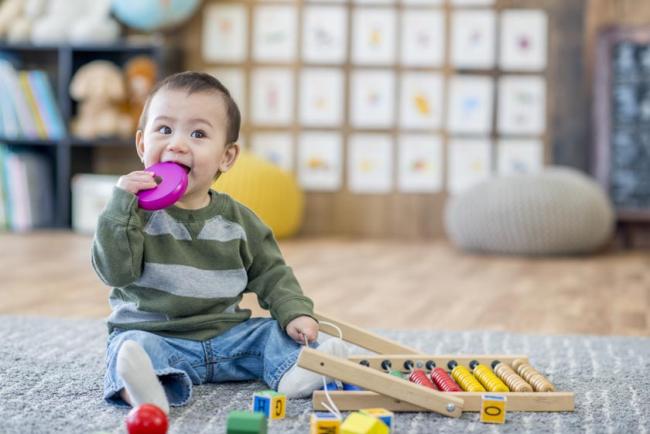 A toddler sitting on a carpet holding a toy in his right hand and chewing another toy in his left hand