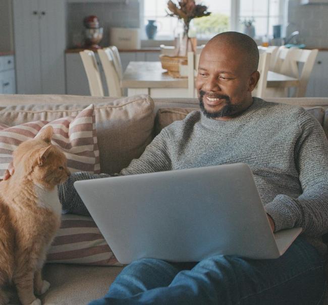 A man sitting on a couch smiling while he pets a cat and works on his laptop. 