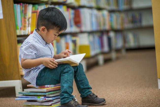 A young boy sitting in a library on a small stack of books while he reads a book