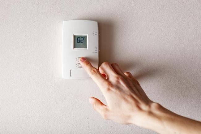 A hand pressing the up button on a house's wall thermostat that displays the temperature of 82 degrees F. 