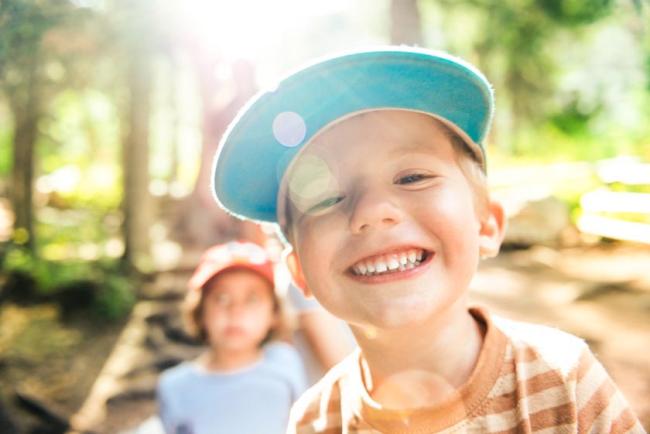 A young boy in a blue hat and striped brown shirt smiles in the foreground as a young girl stands in the background. 